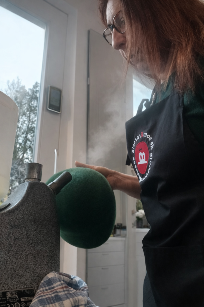 Sombrerera trabajando a mano en la fabricación de un sombrero de fieltro sobre horma de madera en un atelier moderno de sombrerería con herramientas y materiales al fondo.